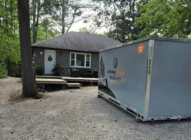 Small gray house with a front porch and wreath, set on a gravel driveway, next to a large gray dumpster labeled with a company name, amid surrounding trees.
